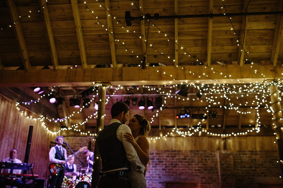 A couple dancing as a band plays behind them inside a barn with fairylights