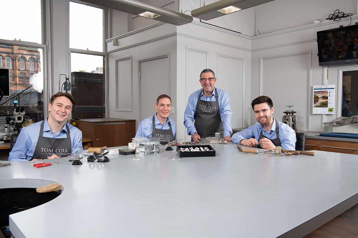 four men in blue collared shirts and grey aprons that say 'Tom Coll Jewellery Workshop' sit around a large workshop space with jewellery and wooden handle tools, smiling at the camera
