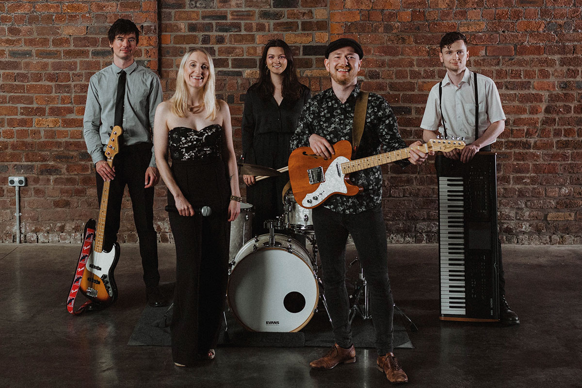 Five-piece wedding band posing with instruments against an exposed brick wall venue backdrop