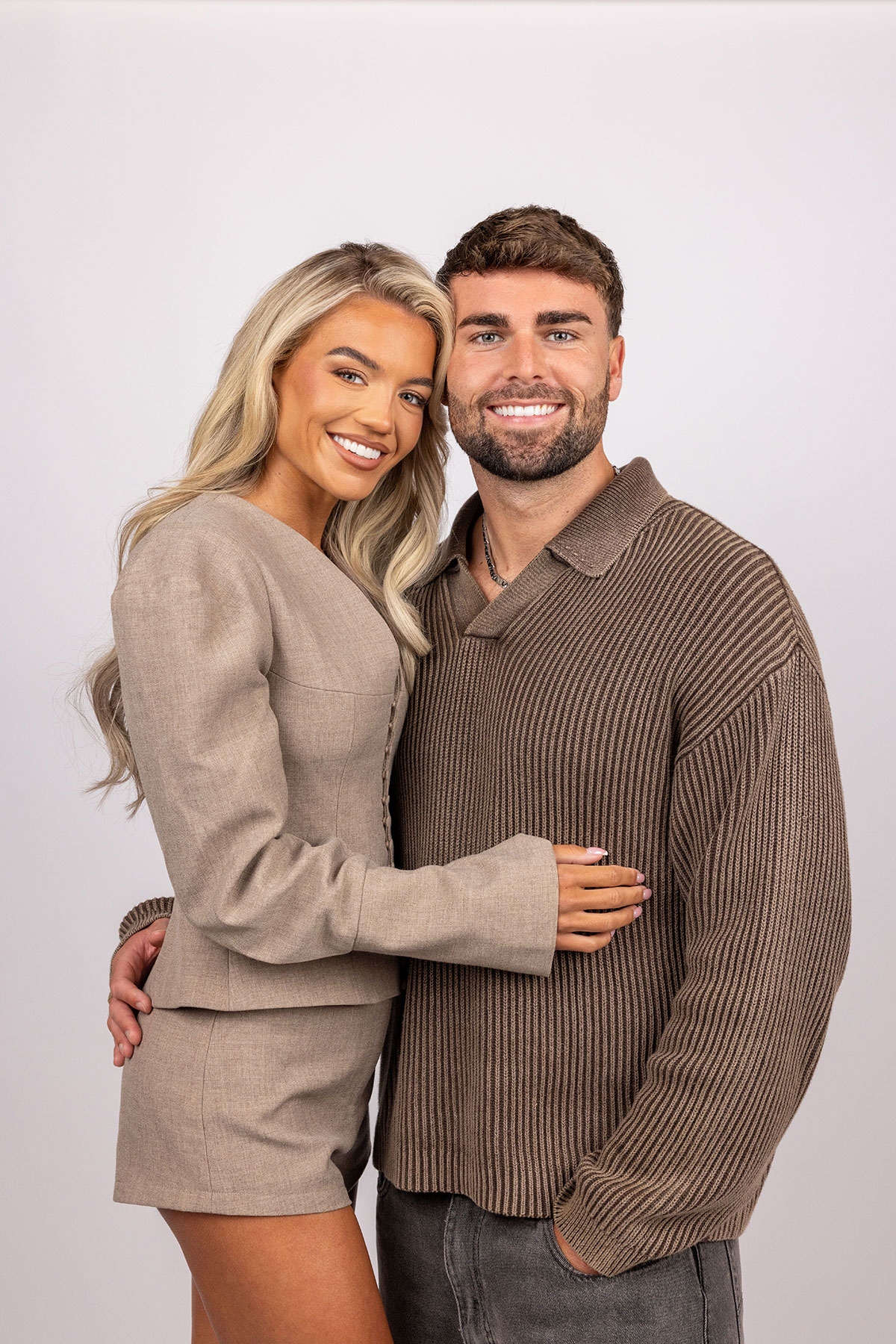 Couple pose in coordinated neutral-toned casual outfits; the woman wears a tailored beige mini dress with long sleeves while the man wears a brown knit jumper, both smiling at the camera