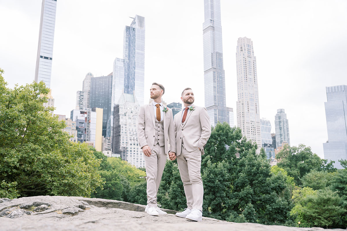 Two grooms holding hands and standing on a rock in Central Park with tall New York skyscrapers rising behind the trees