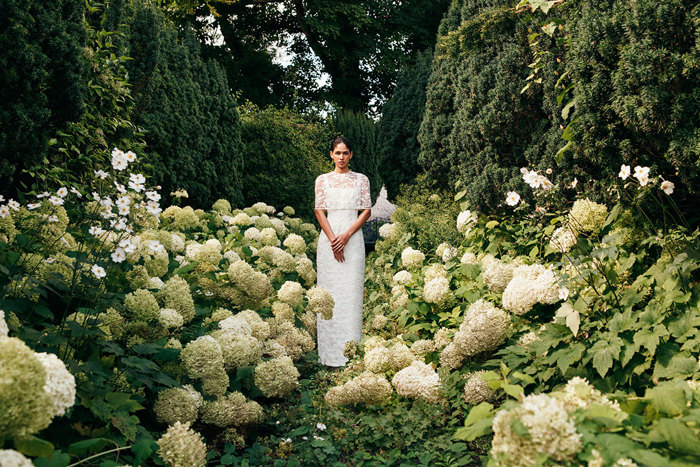 woman in lace wedding dress with lace cape over her shoulders stands in the centre of a green and white flower field with tall hedges around it