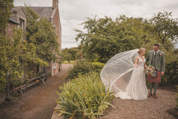 A bride and groom stand in a garden with the bride's veil blowing behind her in the wind