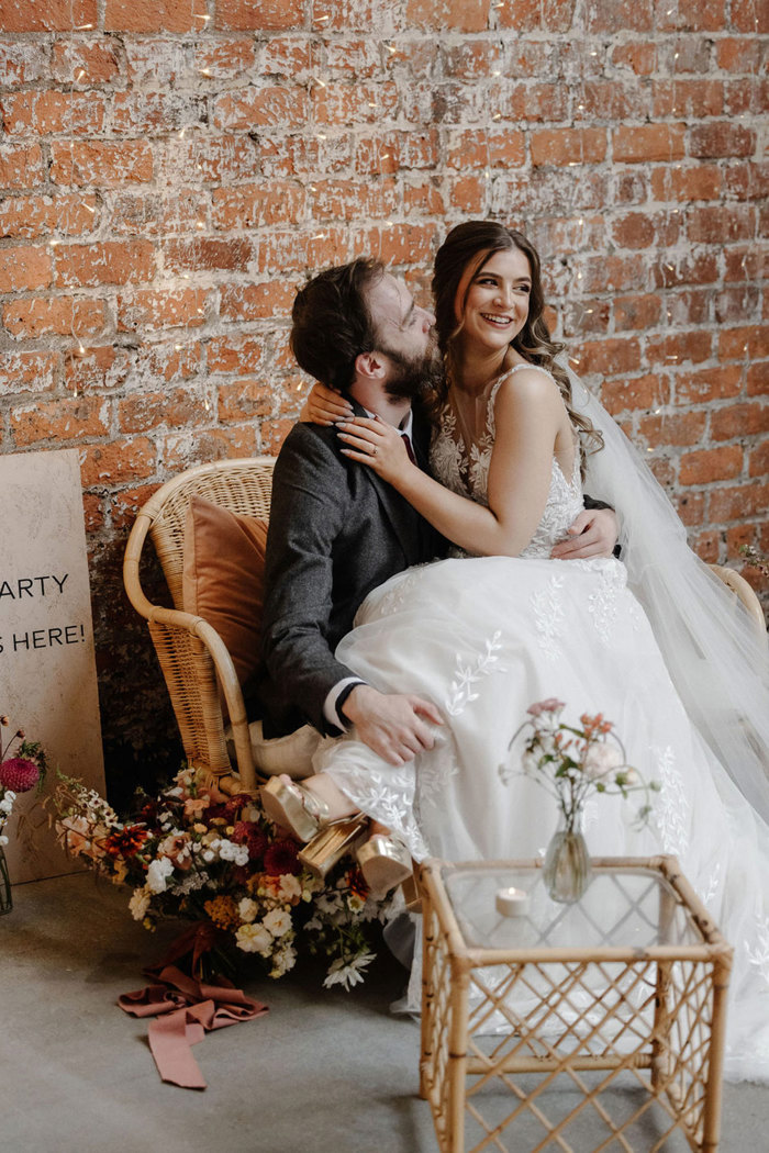 bride sits smiling on groom's lap with her arms wrapped around his neck and a large bouquet of wildflowers at her feet