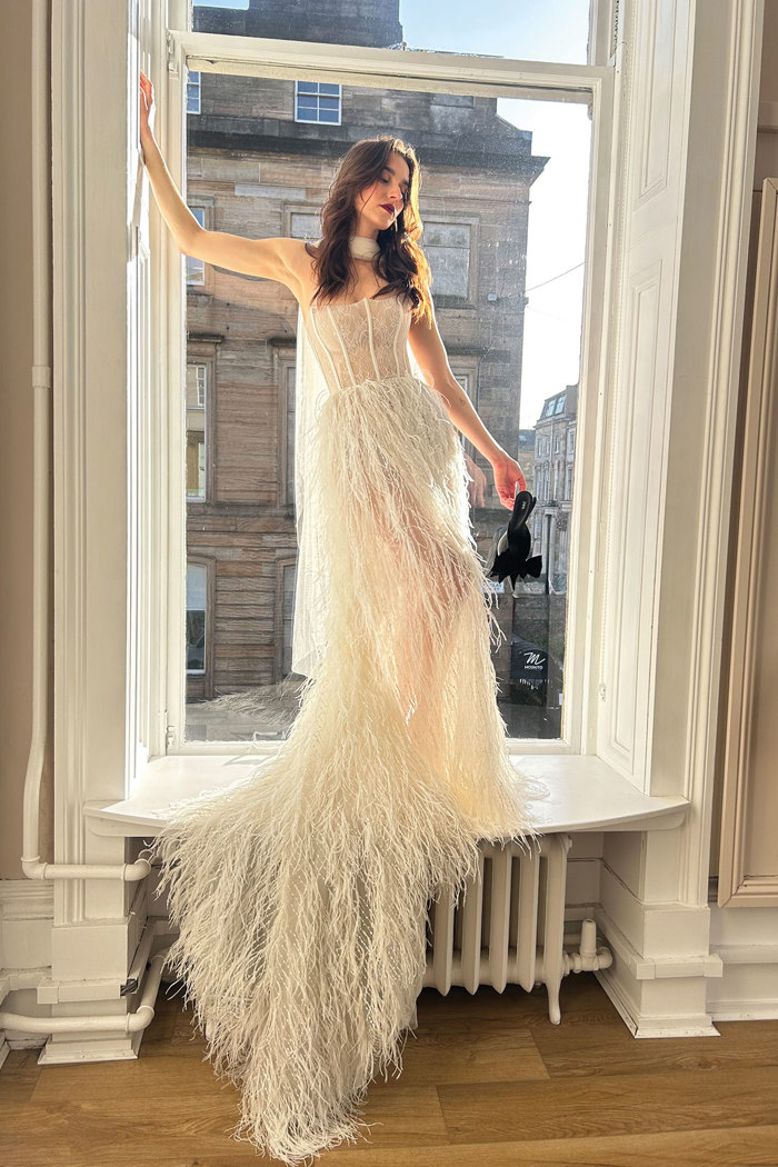 brunette woman in strapless feather skirt wedding dress and neck scarf stands on sill of large window, holding a single black heel in one hand