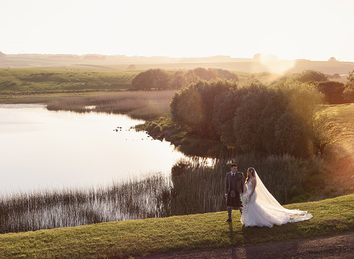 a bride and groom walking across grass at sunset with lake and bushes in background