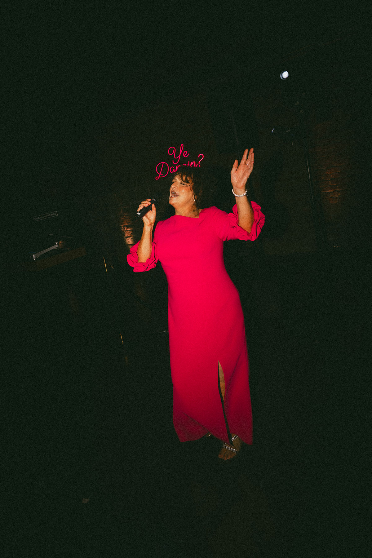 Woman in a bright pink dress sings into a microphone against a dark background, one hand raised, wearing a large matching fascinator