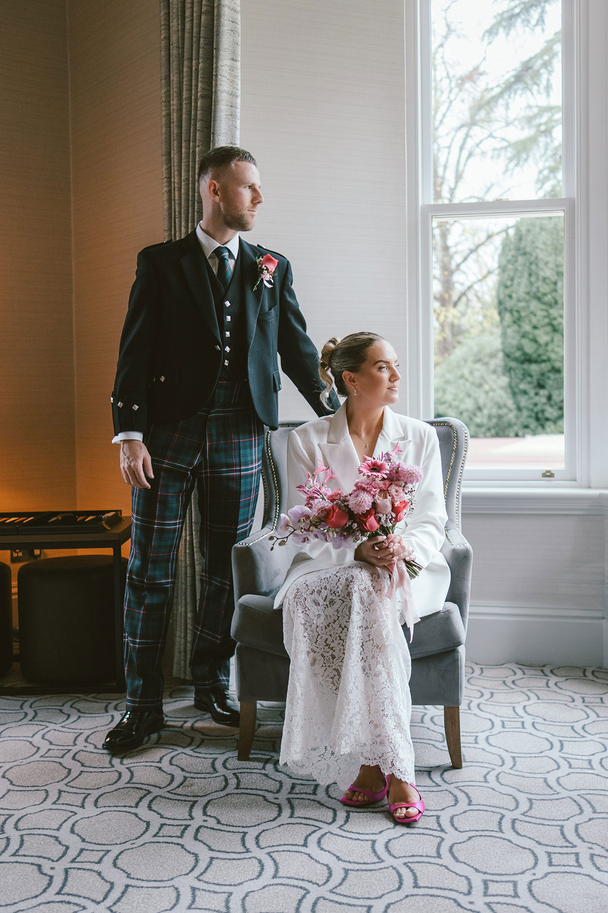 Bride seated with bouquet by window and groom standing behind in natural light wedding portrait