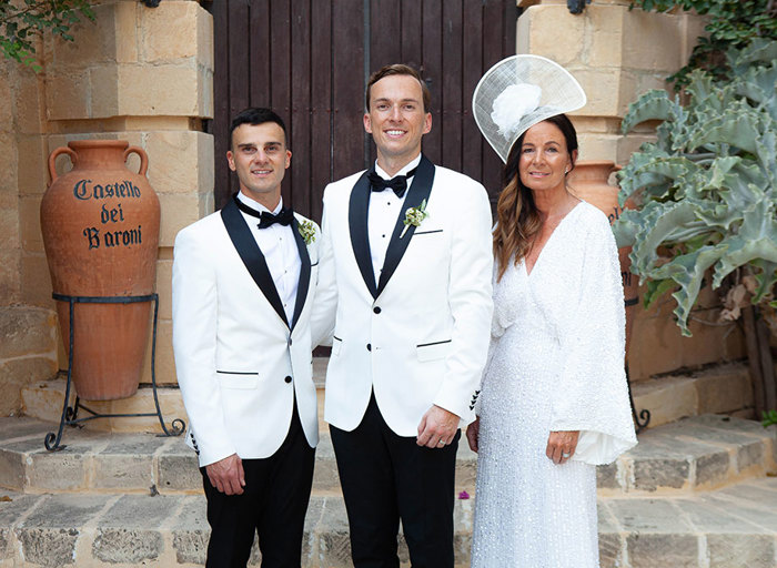 two grooms posing with a woman wearing a white embellished dress in front of an old stone building with large wooden door