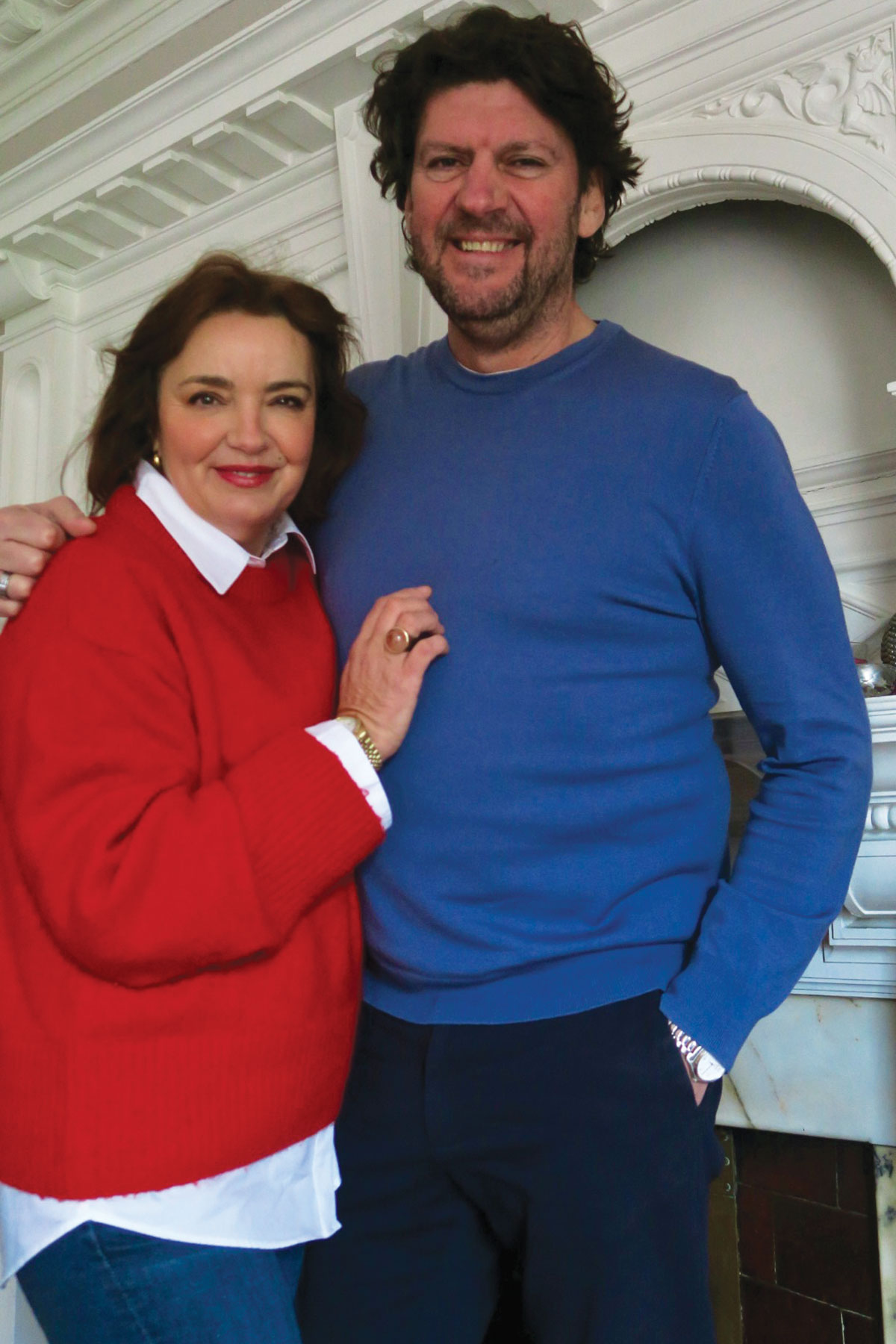 Couple standing indoors in front of a white fireplace; the woman wears a red jumper layered over a white shirt and the man wears a blue sweater