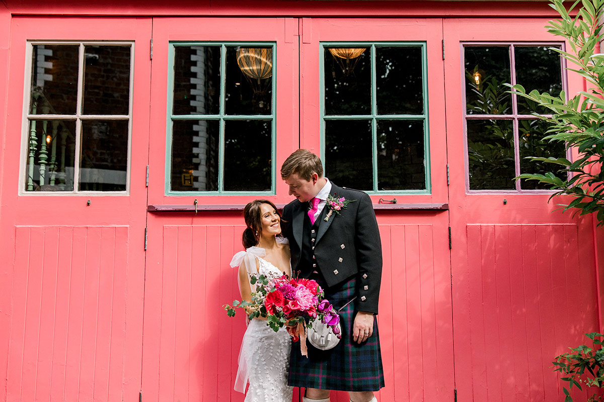 Bride and groom posing in front of vibrant pink wooden doors with a colourful bouquet