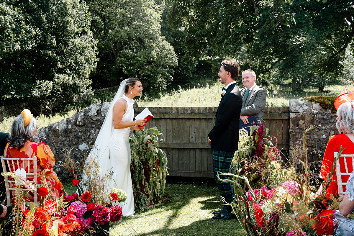 Bride reads her vows during an outdoor orchard ceremony at Kinkell Byre, surrounded by vibrant red, pink and orange flowers at this colourful Fife barn wedding.