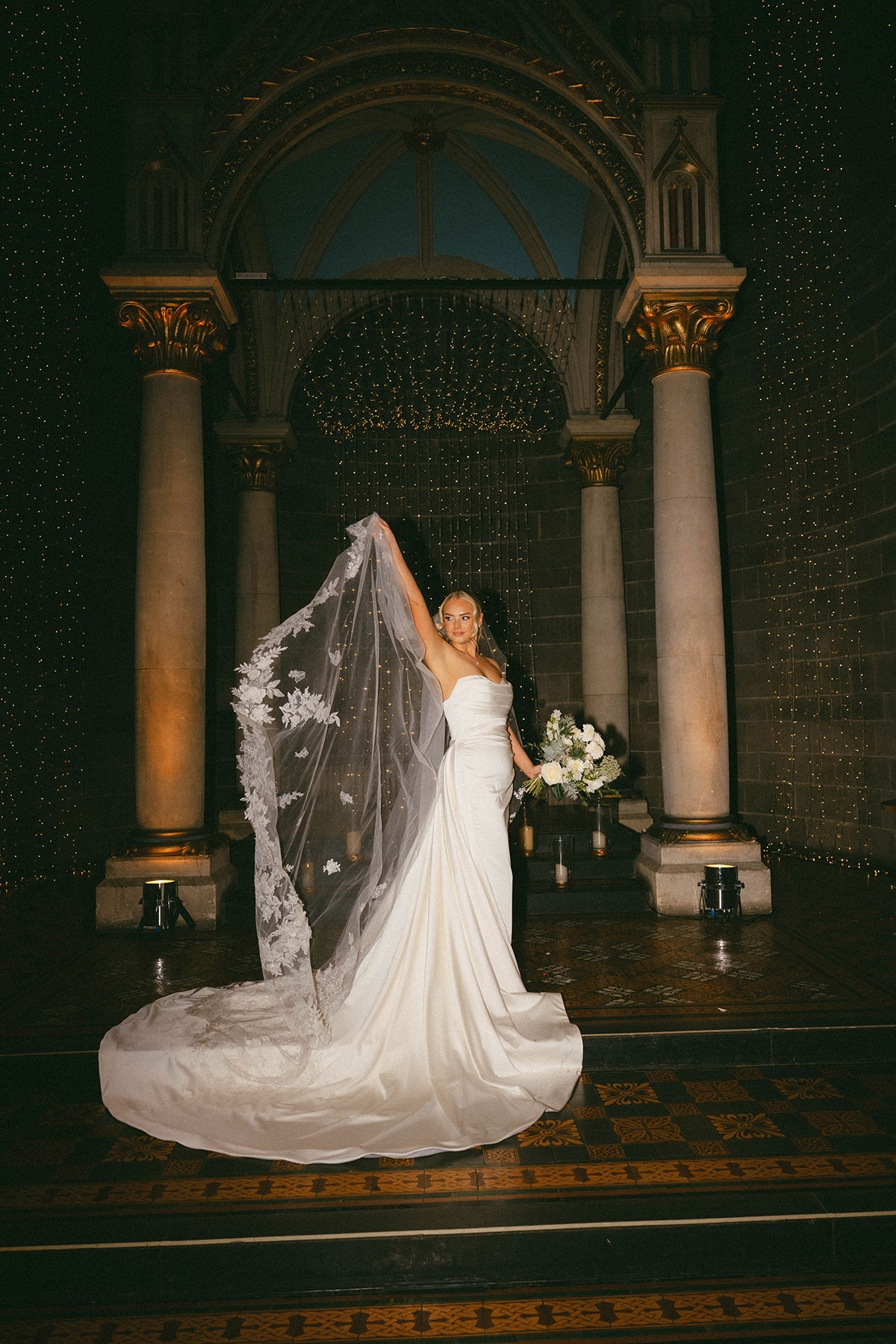 bride posing with veil and bouquet under ornate arch with warm lighting