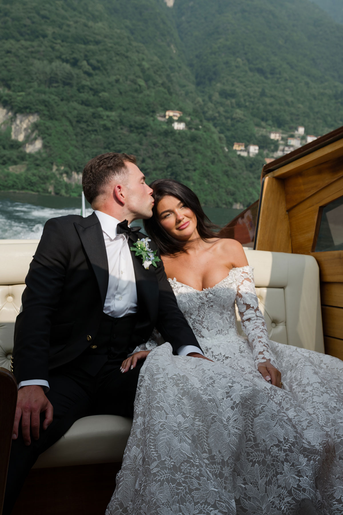 Bride and groom relaxing on a wooden boat on Lake Como, with green hills and lakeside villages in the background.