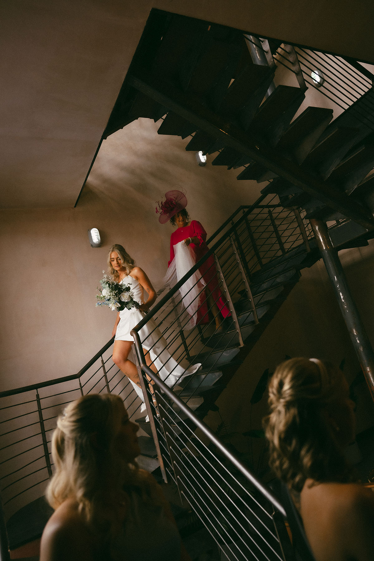 Bride in a white gown holding a bouquet walks down a modern staircase followed by an older woman in a bright pink dress and fascinator, with other guests visible below