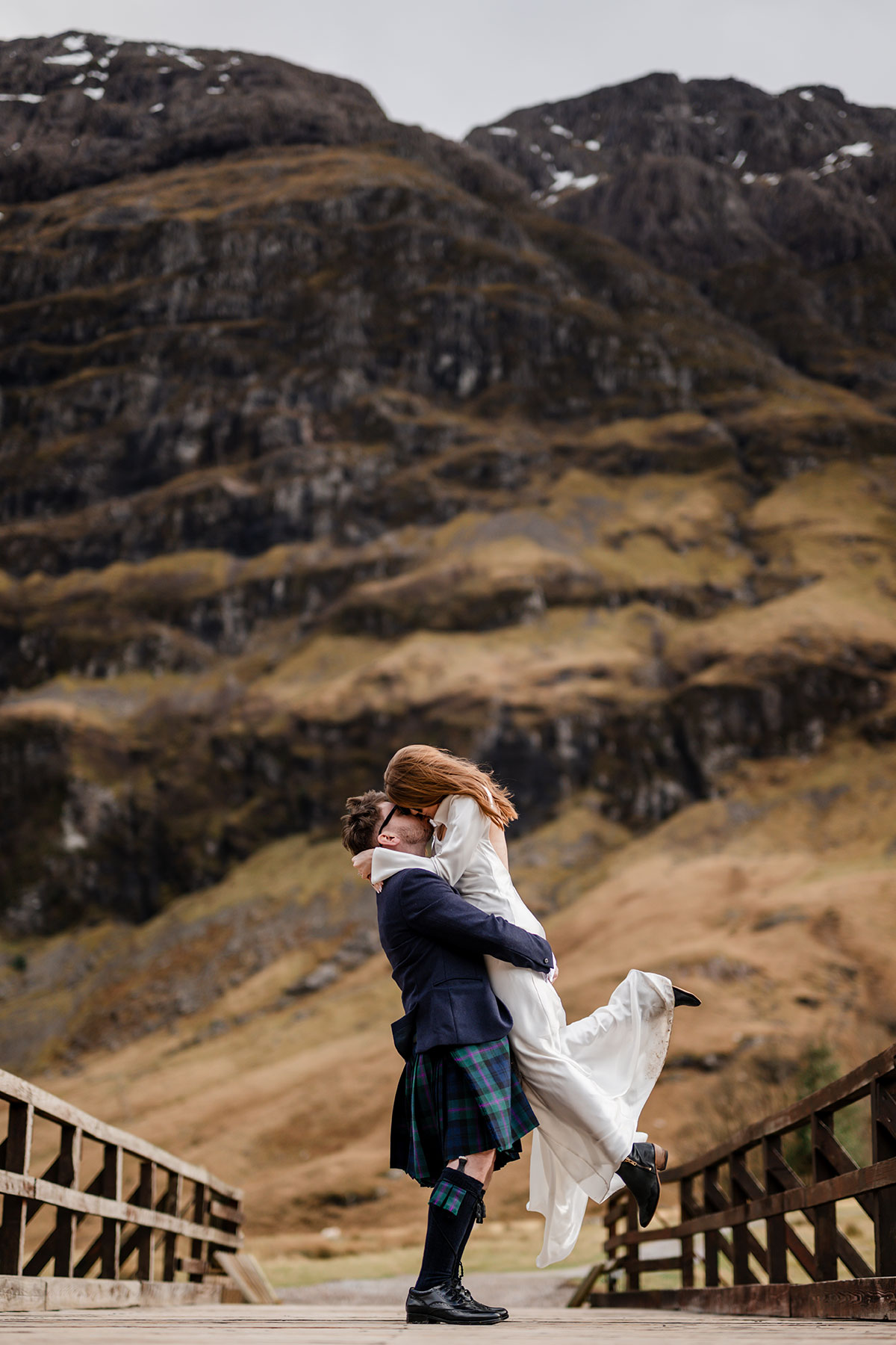 Bride and groom celebrating with a joyful lift and kiss on a bridge in Glencoe after their Scottish elopement ceremony