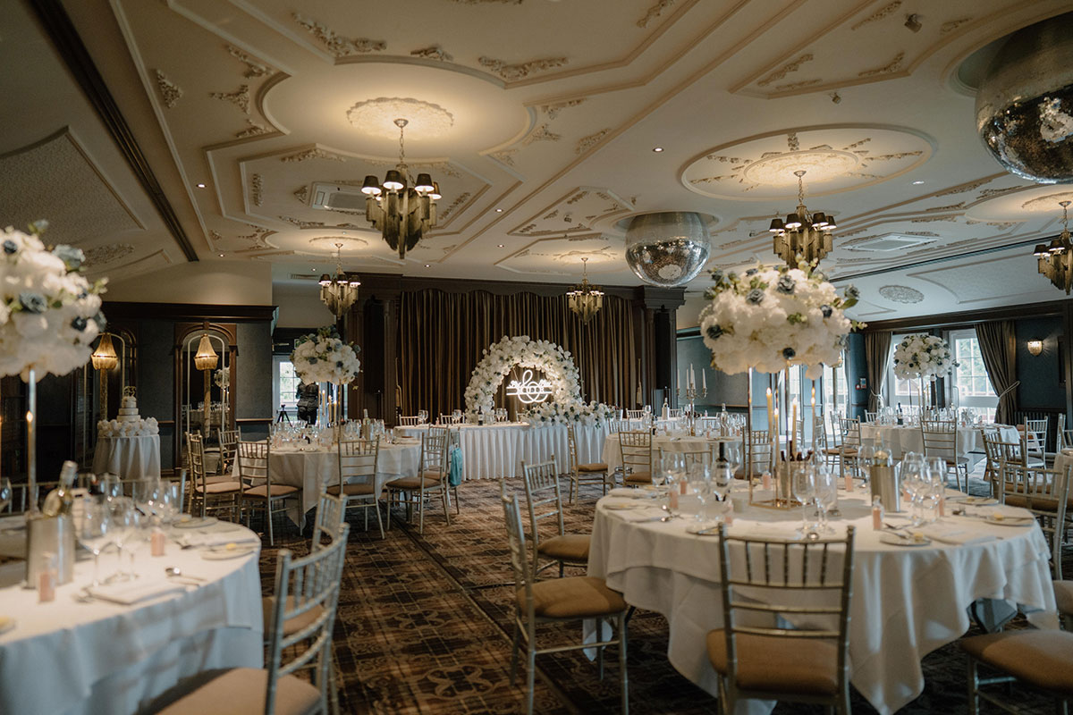 Wedding reception setup inside Cornhill Castle, featuring round tables, floral centrepieces, mirrored décor and a white floral backdrop beneath ornate ceilings
