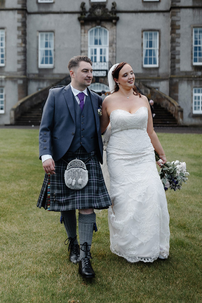 groom in full tartan outfit and bride in strapless floral wedding dress with long veil and bouquet walk in grass in front of large stone building