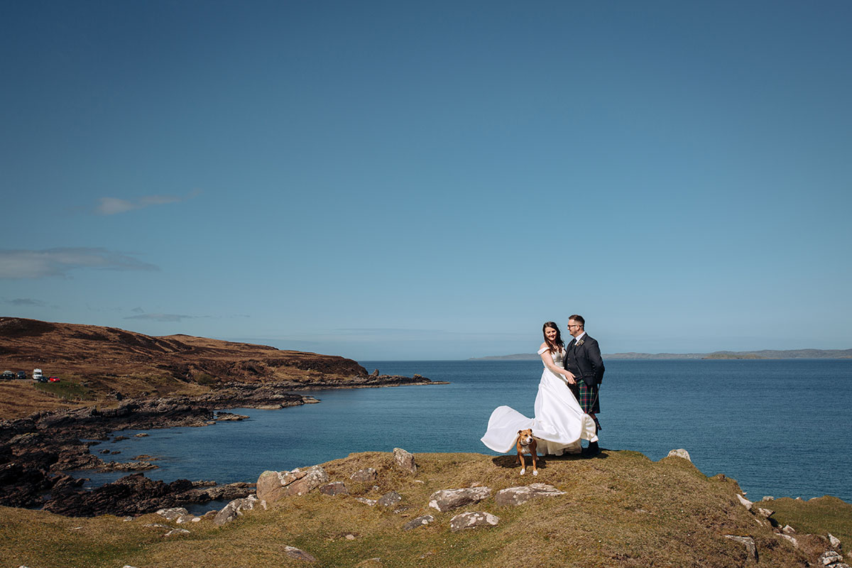 Bride and groom stand on a grassy cliff with panoramic views of the Scottish coastline at Achnahaird Bay.