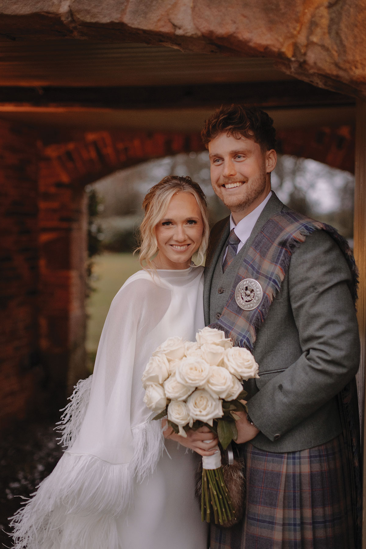 blonde bride in feathery cape and white wedding dress holds bouquet of white roses and stands beside groom in green jacket and red, blue, green kilt with matching fly plaid
