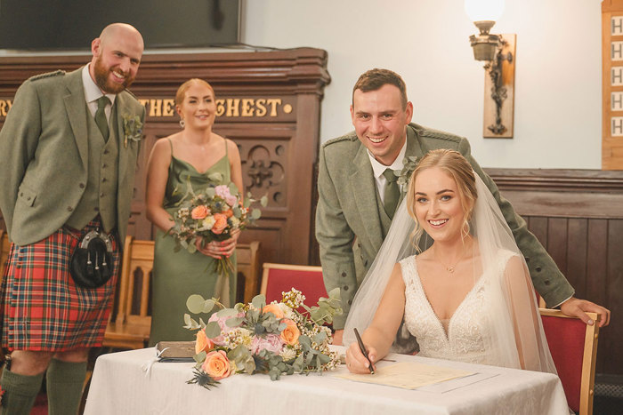 A bride sitting down signing the marriage license while the groom, a bridesmaid and a groomsmen stand behind her
