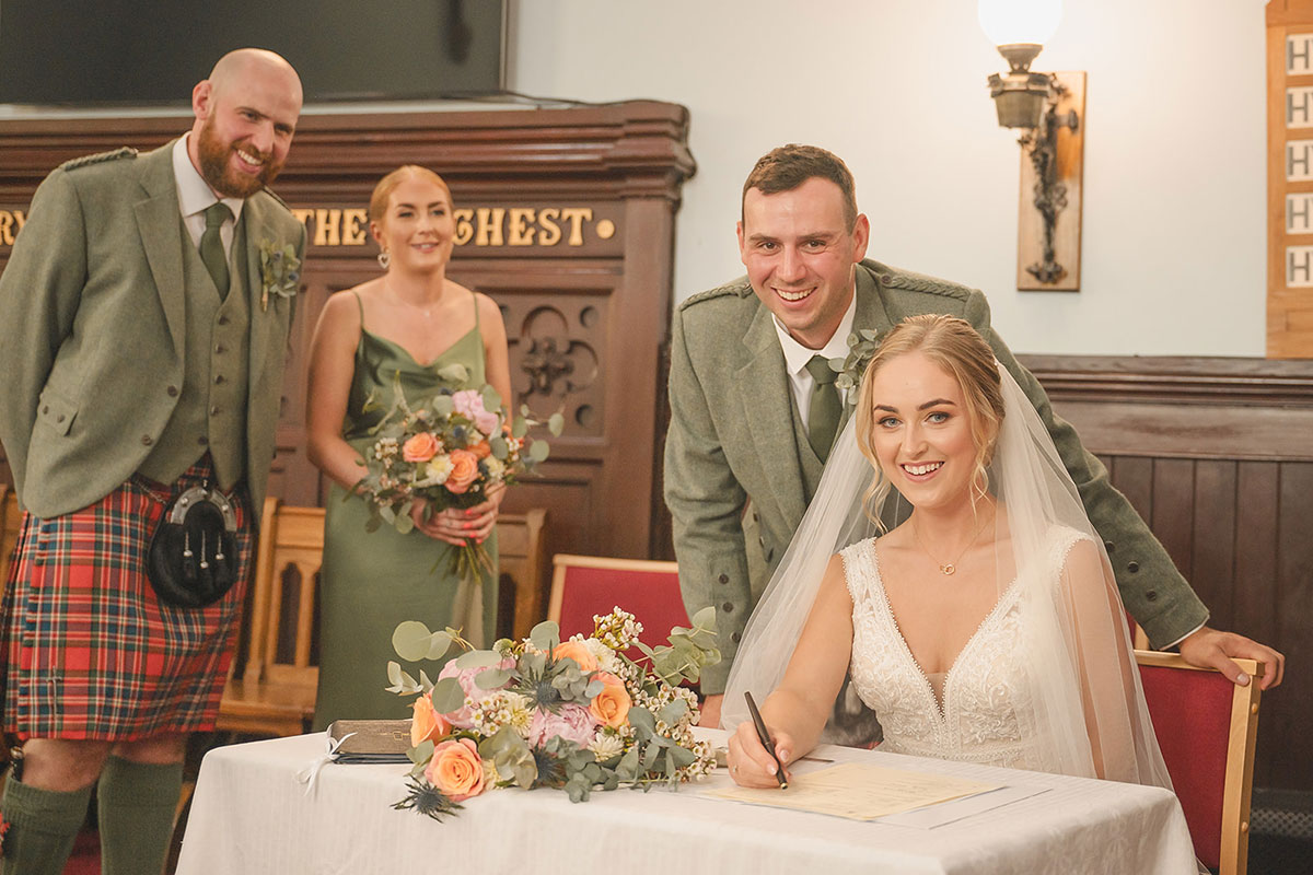 A bride sitting down signing the marriage license while the groom, a bridesmaid and a groomsmen stand behind her