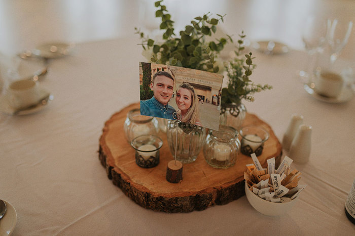 A Log Slice Centrepiece On A White Tablecloth With Photo Surrounded By Greenery And Tea Lights