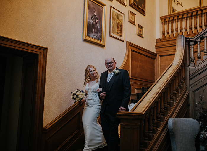 A woman in a white dress and a man in a suit walk down a wooden staircase arm in arm