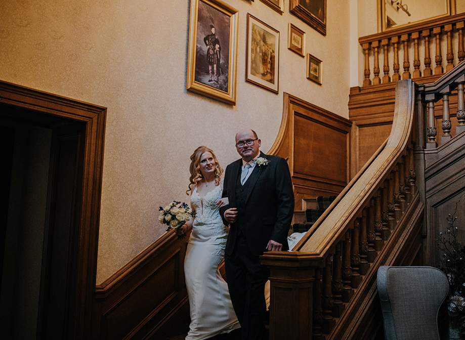 A woman in a white dress and a man in a suit walk down a wooden staircase arm in arm