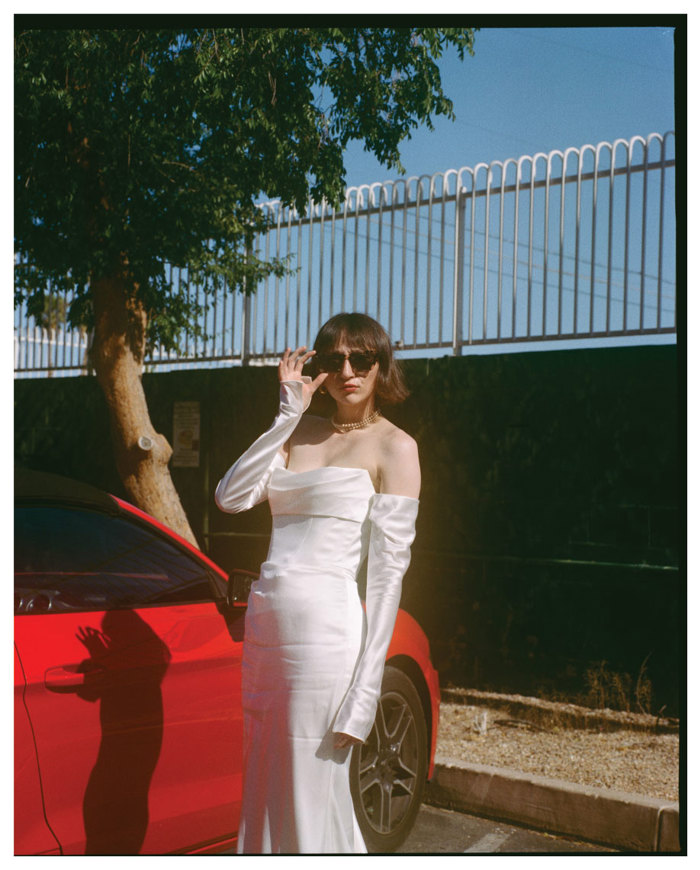 a bride fixing her sunglasses while standing next to a red car. She is wearing a long white dress with removable sleeves