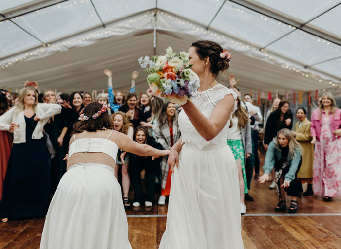 one bride wearing a fine lace top and flowing skirt prepares to throw a colourful bouquet as a second bride encourages a small crowd of excited-looking wedding guests to participate. They are in marquee setting with wooden floor, colourful streamers hanging from the ceiling and fairy lights