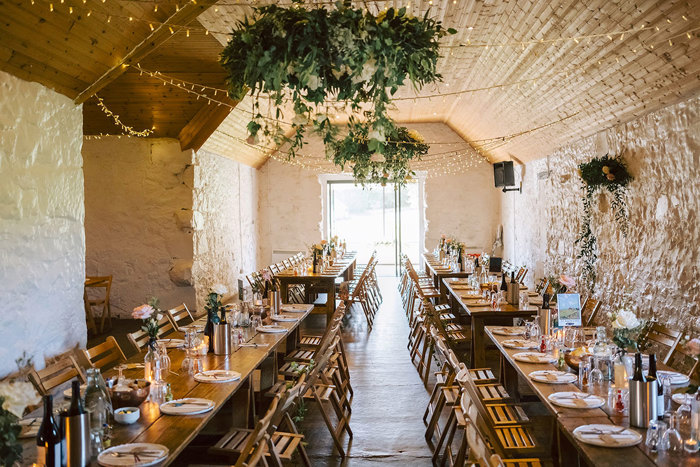 The inside of a stone barn with long tables set out for a meal and foliage and fairylights hanging from the ceiling 