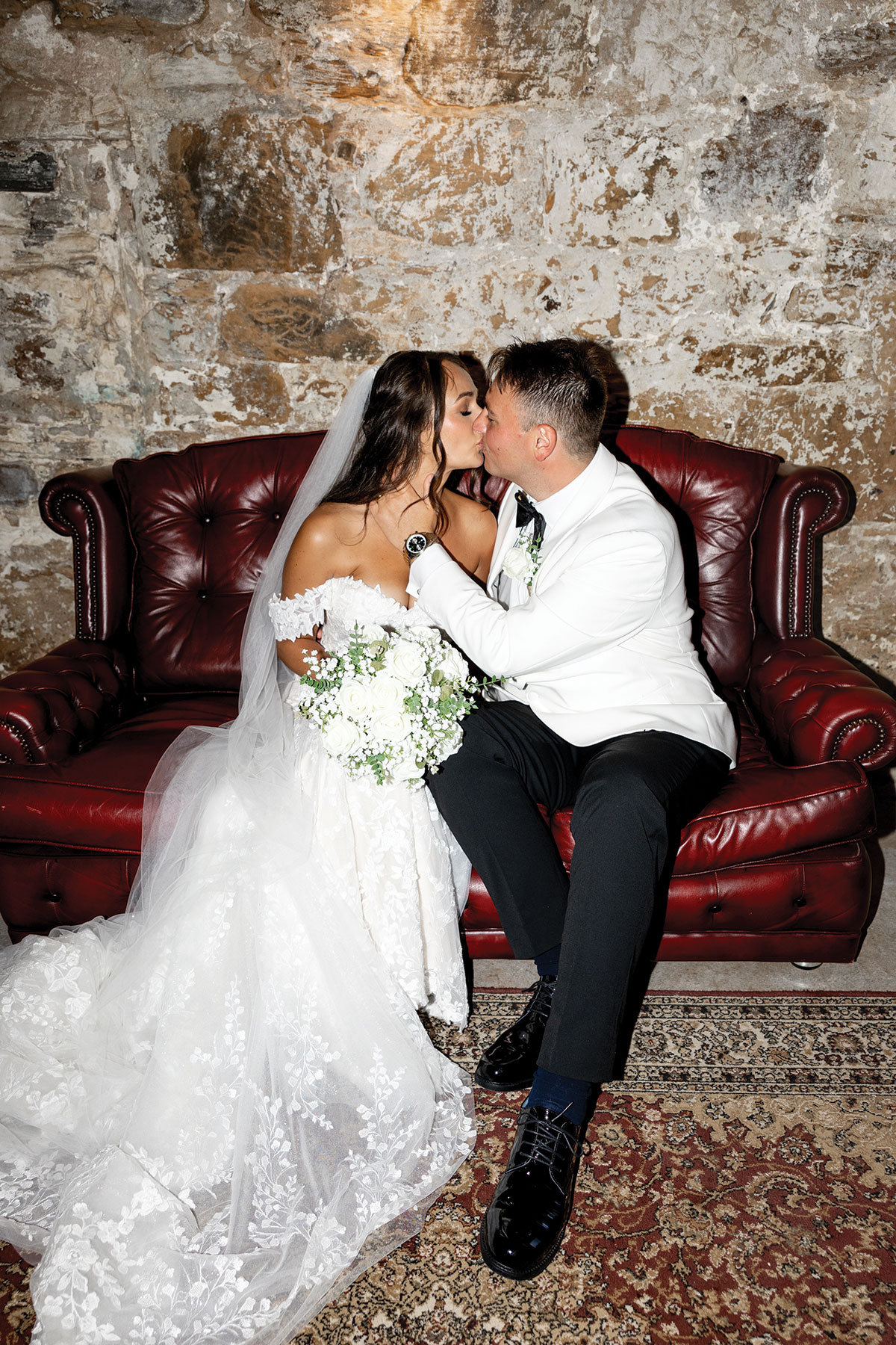 The couple sit on a red leather sofa inside Falside Mill, sharing a kiss against the textured stone wall backdrop.