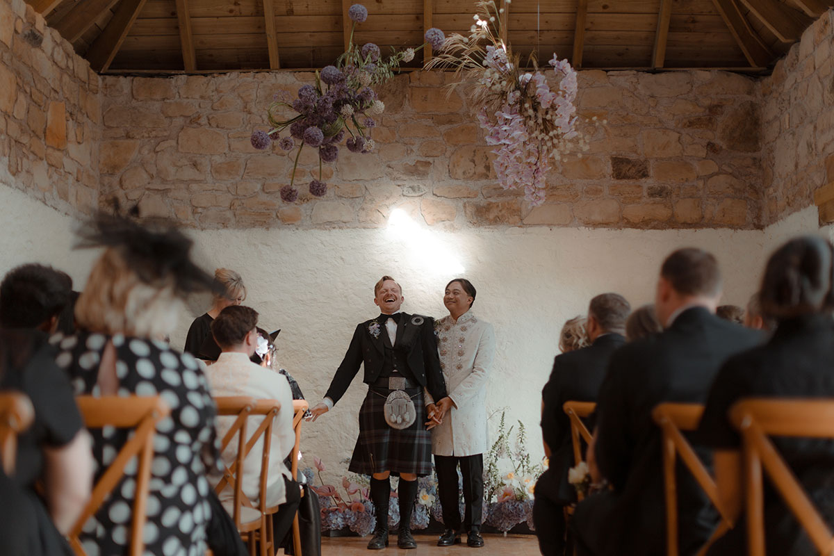 Two grooms holding hands during an intimate barn wedding ceremony at their renovated farm near Falkirk under a flower cloud