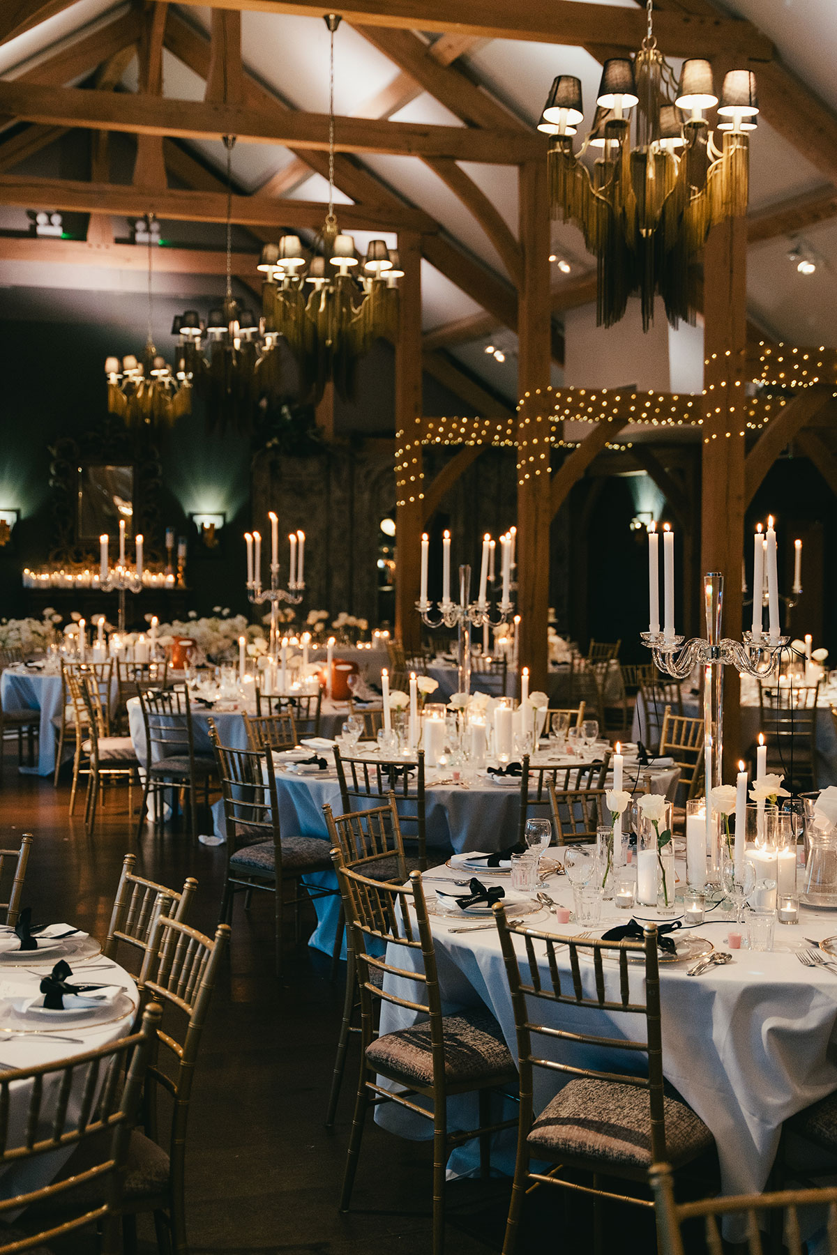 Wedding reception setup with round tables, tall candelabras, fairy lights and chandeliers in rustic barn at Enterkine Country House Ayrshire