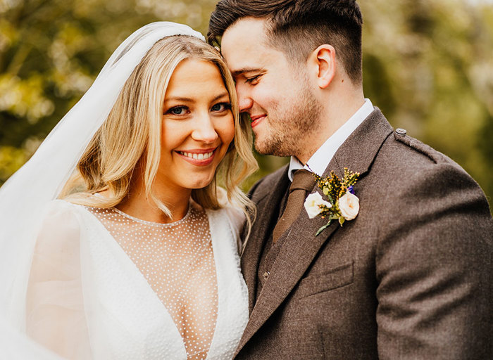 a close up portrait of a smiling bride and groom. The bride is wearing a dotted tulle dress and veil and the groom is wearing a brown jacket. He is nuzzling into the side of the bride's face