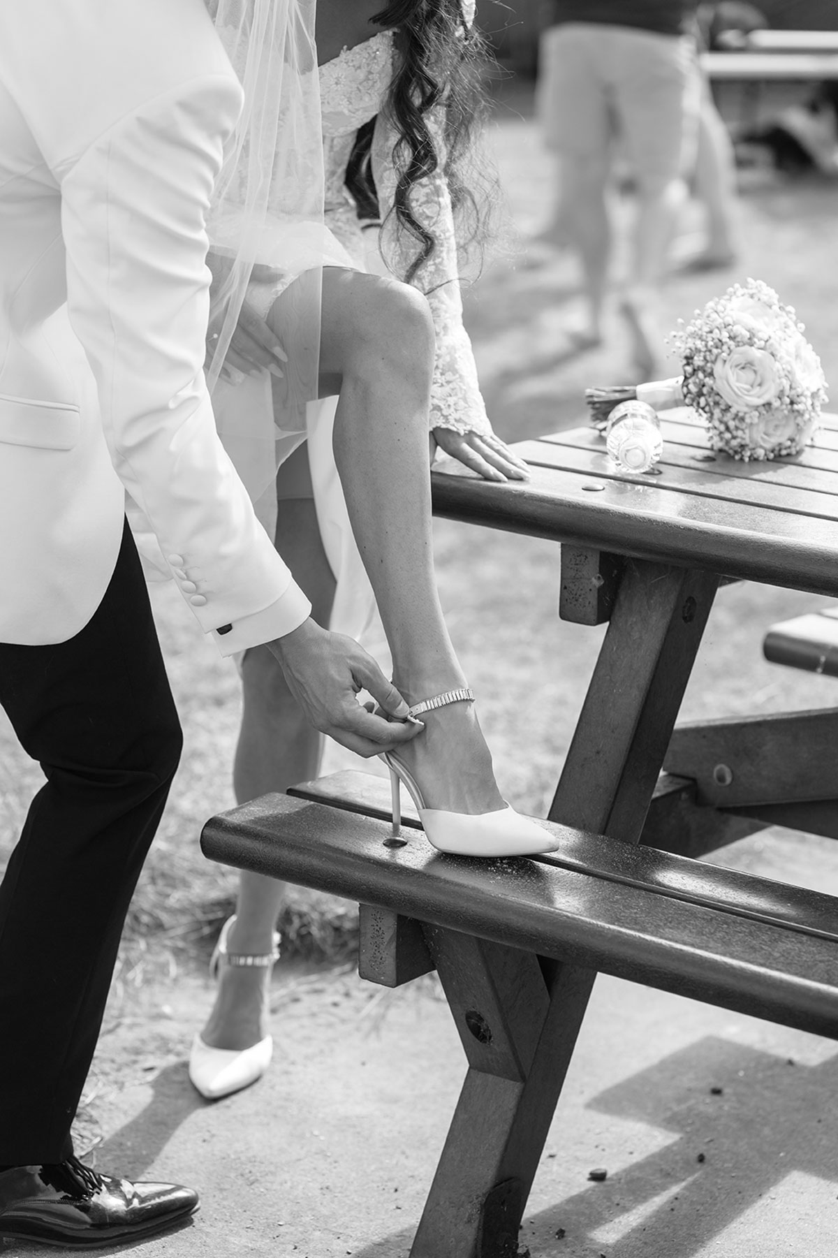 Groom fastening the bride’s white heel strap during wedding preparations, black-and-white detail