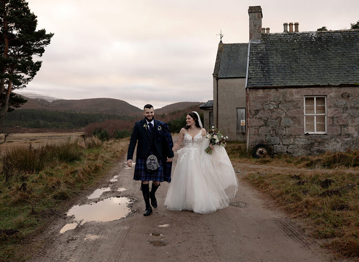 a laughing bride and groom walking on a path in front of an old stone cottage. There is snow on distant hills and the surrounding landscape looks brown and autumnal