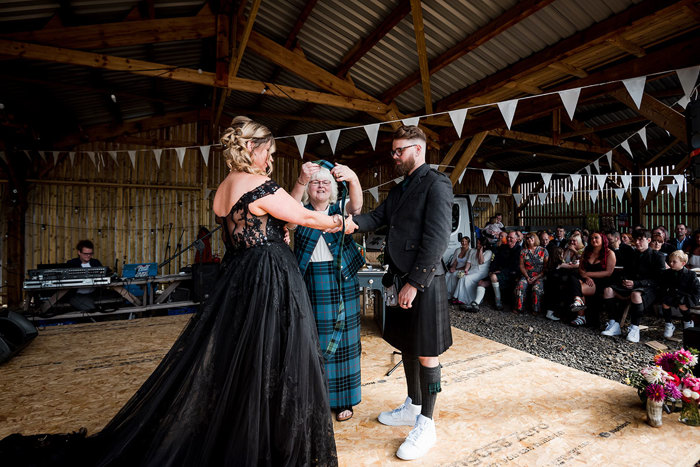 A person placing a tartan ribbon over the hands of a bride and groom wearing black during a wedding ceremony in a wooden shed building