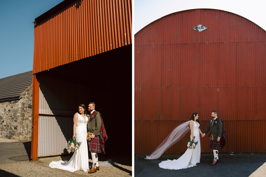 A bride in a white dress and a groom in a red kilt pose for photos in front of a red barn