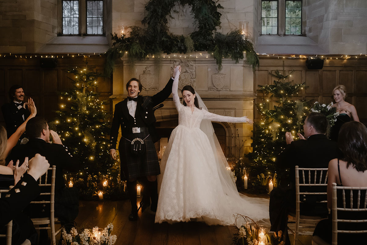 a cheering bride and groom standing in front of a large stone fireplace that's illuminated by fairy lights and has Christmas trees either side.