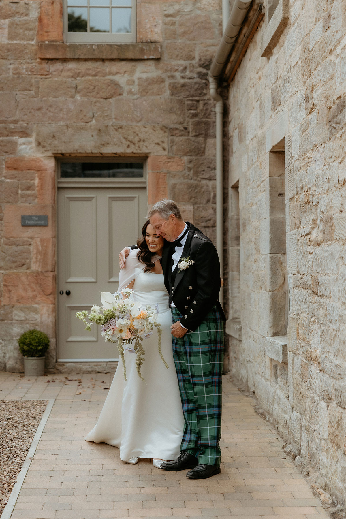 Bride and father embracing in courtyard of Rosebery House and Steading, Midlothian, ahead of chapel wedding ceremony.