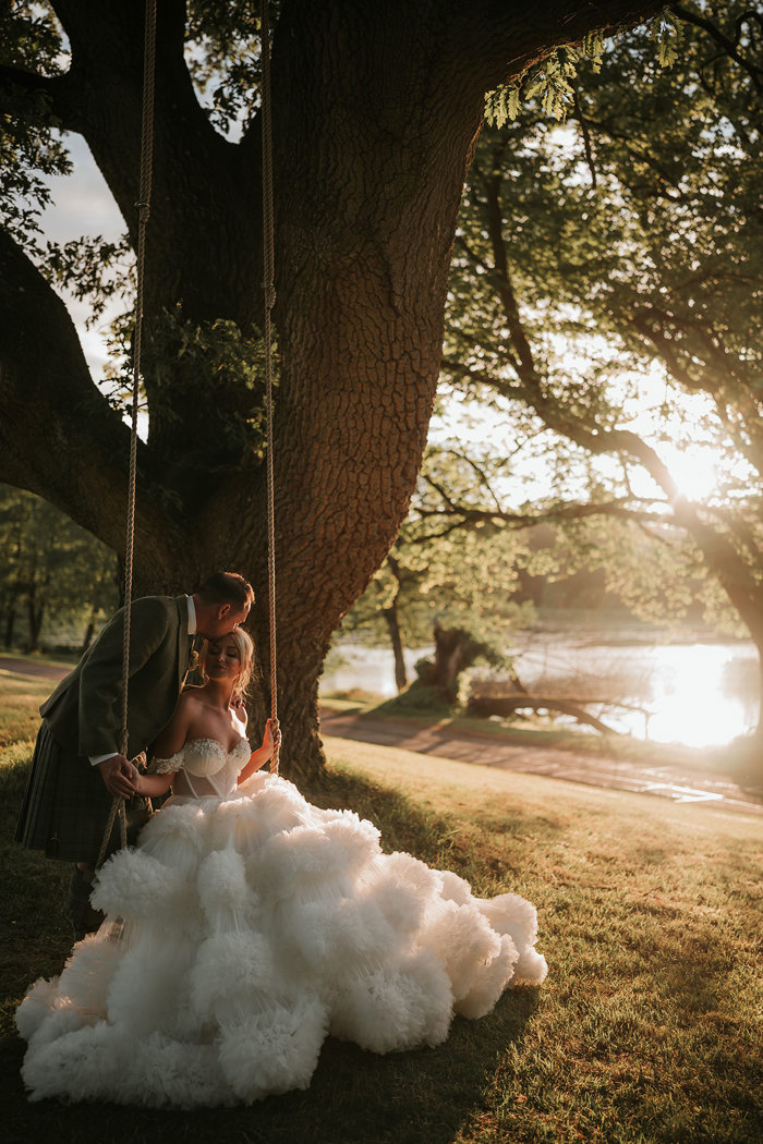 A bride wearing a wedding dress with a voluminous tulle skirt sitting on swing with a groom in a green kilt standing behind her and kissing her forehead