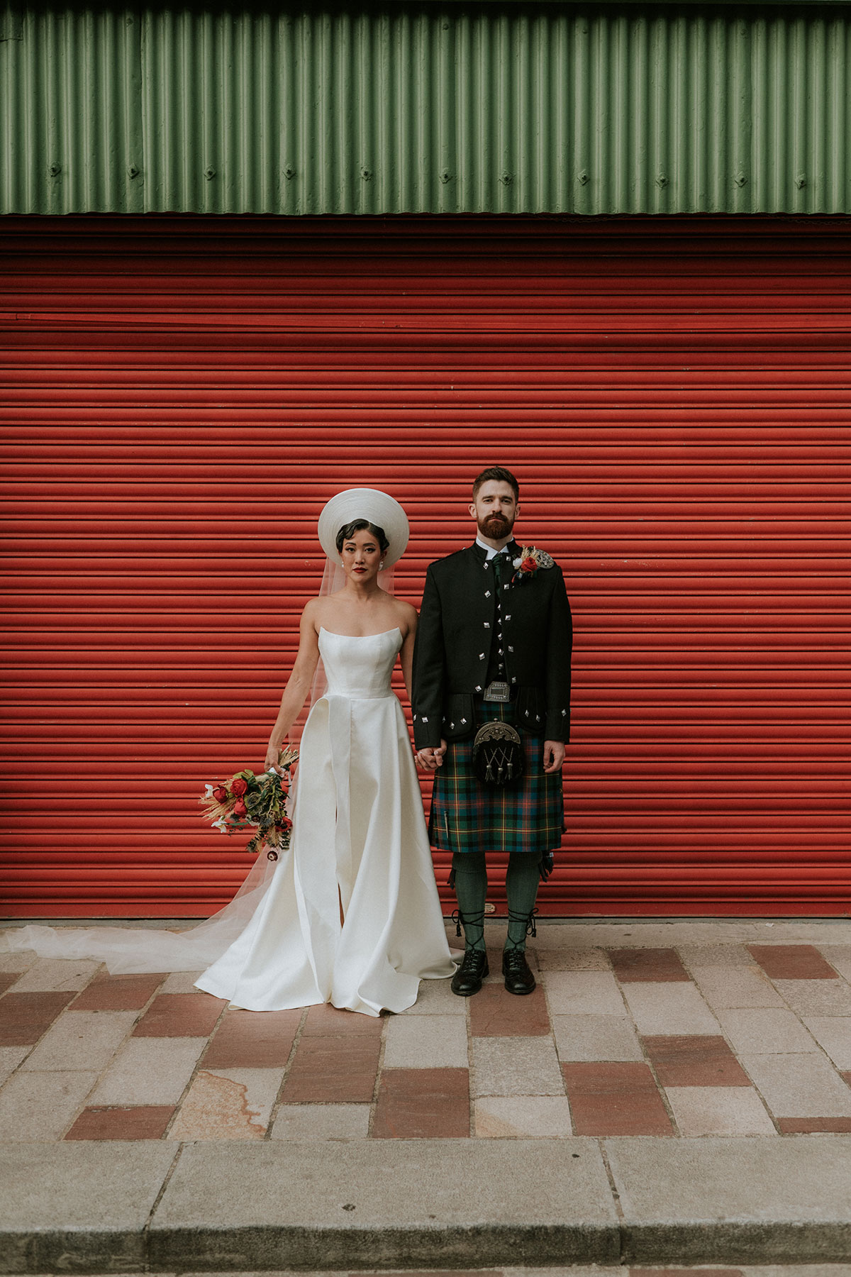 Bride and groom holding hands in front of red shutter at Barras Art and Design Glasgow wedding venue