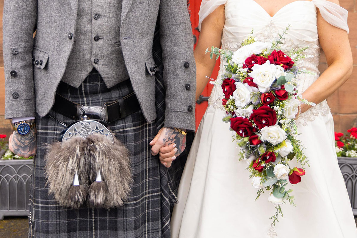 groom in grey tartan kilt and bride with red and white bouquet are photographed torso down and holding hands