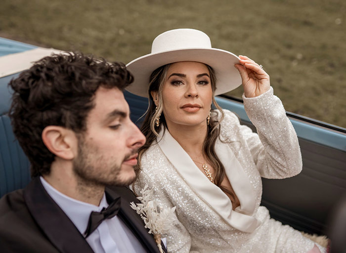 A close up photo of a man and a woman in a car, the man looks ahead as the woman touches the brim of her hat and looks at the camera