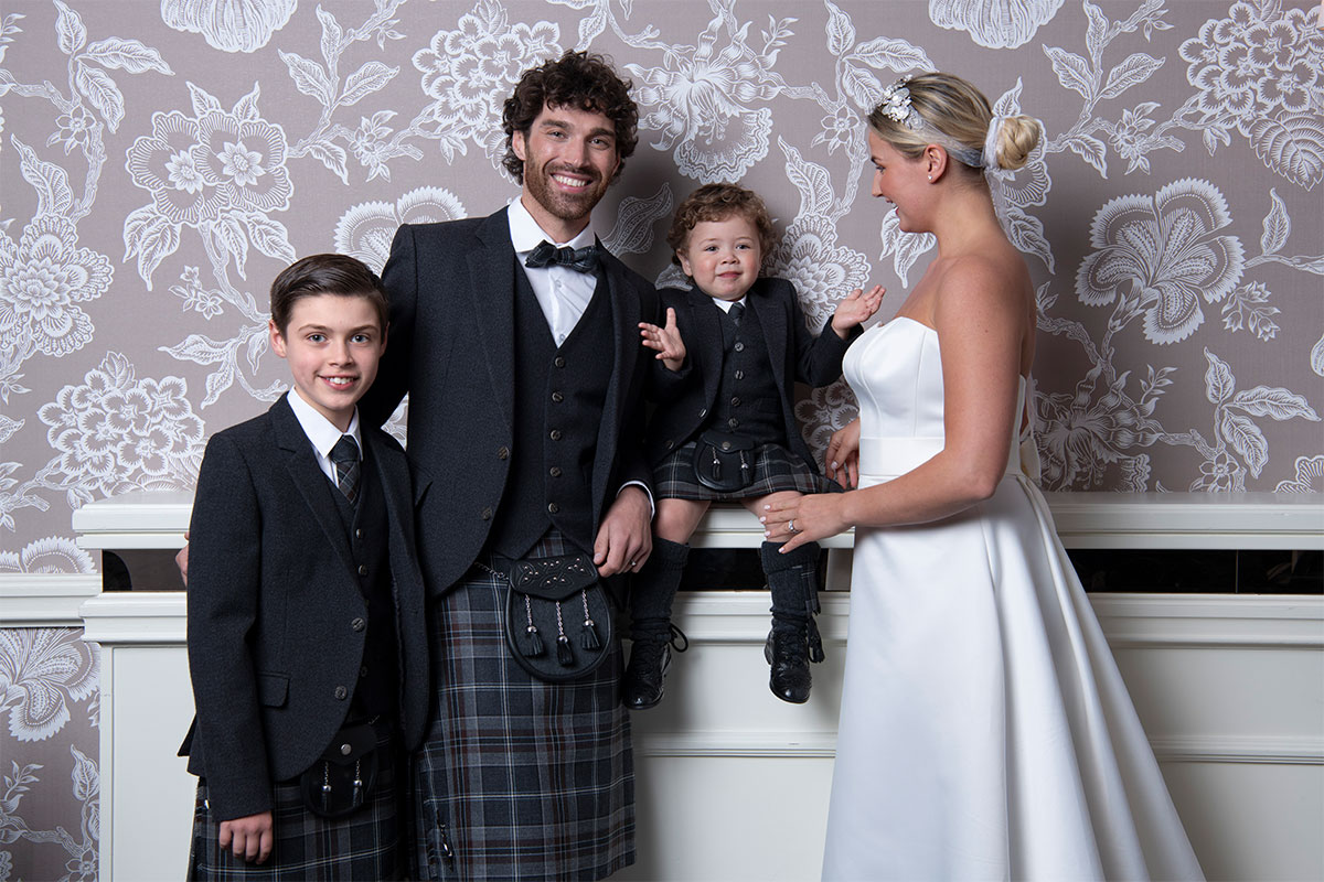 Smiling man wearing highlandwear with bride standing in side profile, next to young boy in highlandwear and clapping toddler sitting on ledge in highlandwear