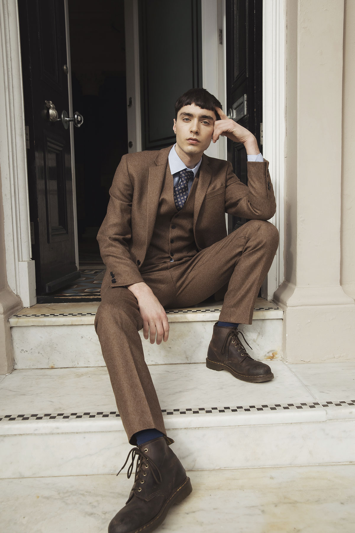 man with short dark hair sits on white marble steps in front of black door wearing brown suit set, brown tie-up boots, a pale blue collared shirt and a dark blue and red spotted tie