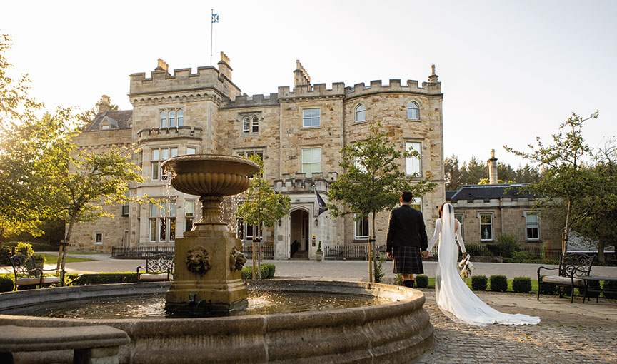 Bride and groom holding hands in front of Crossbasket Castle, framed by the courtyard fountain.