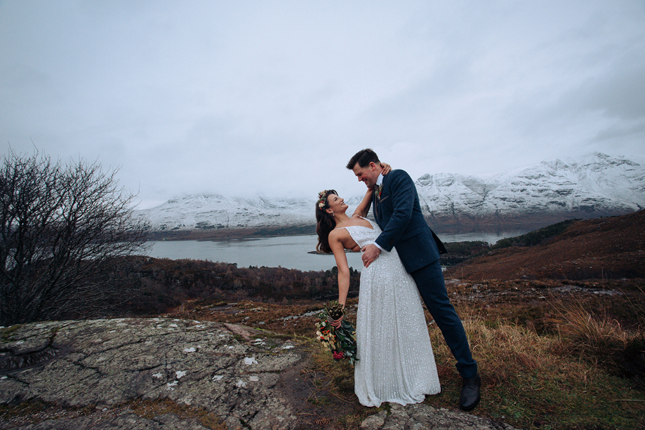 Bride and groom look at each other in front of loch with snow-capped mountains in the background